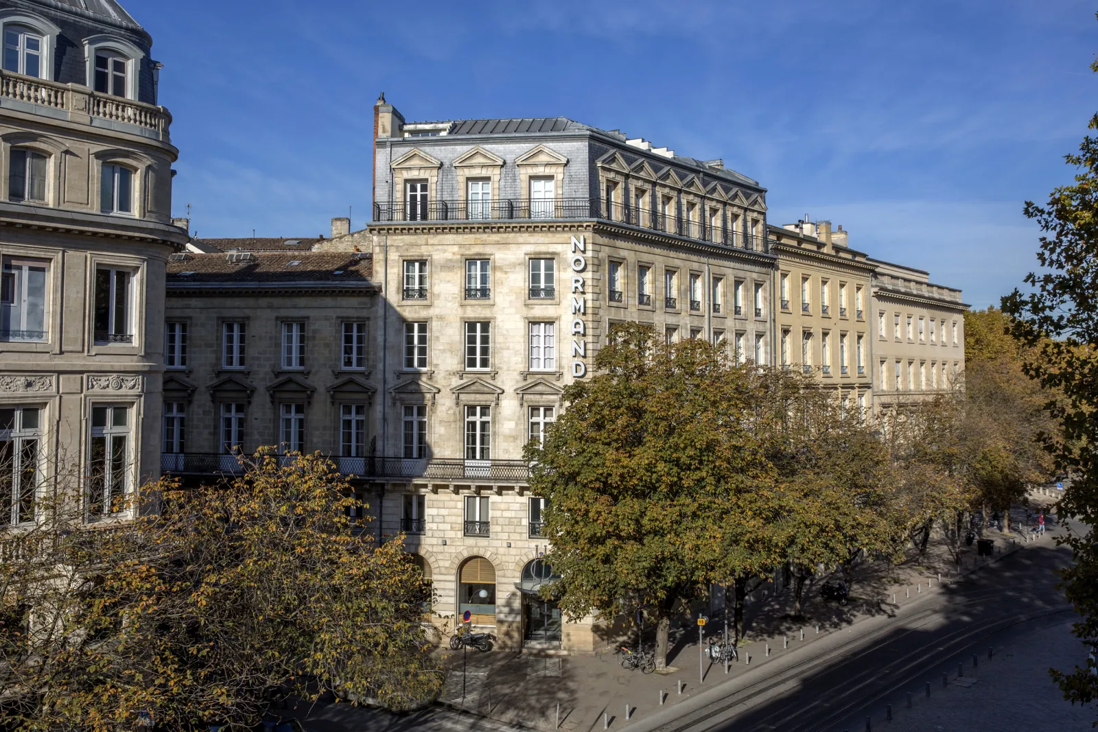 Facade of the Hôtel de Normandie in Bordeaux, a historic Haussmann-style building featuring mansard roofs and architectural ornaments, surrounded by autumn foliage.