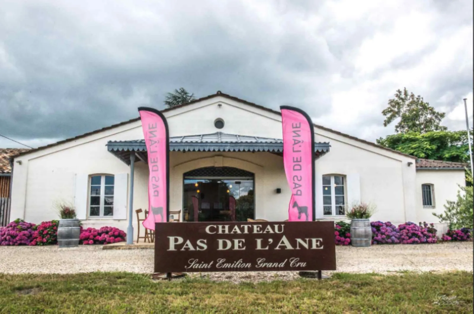 White facade of Château Pas de l'Ane, a Saint-Émilion Grand Cru winery in Bordeaux, featuring pink banners, magenta flowers, and a brown wooden sign.