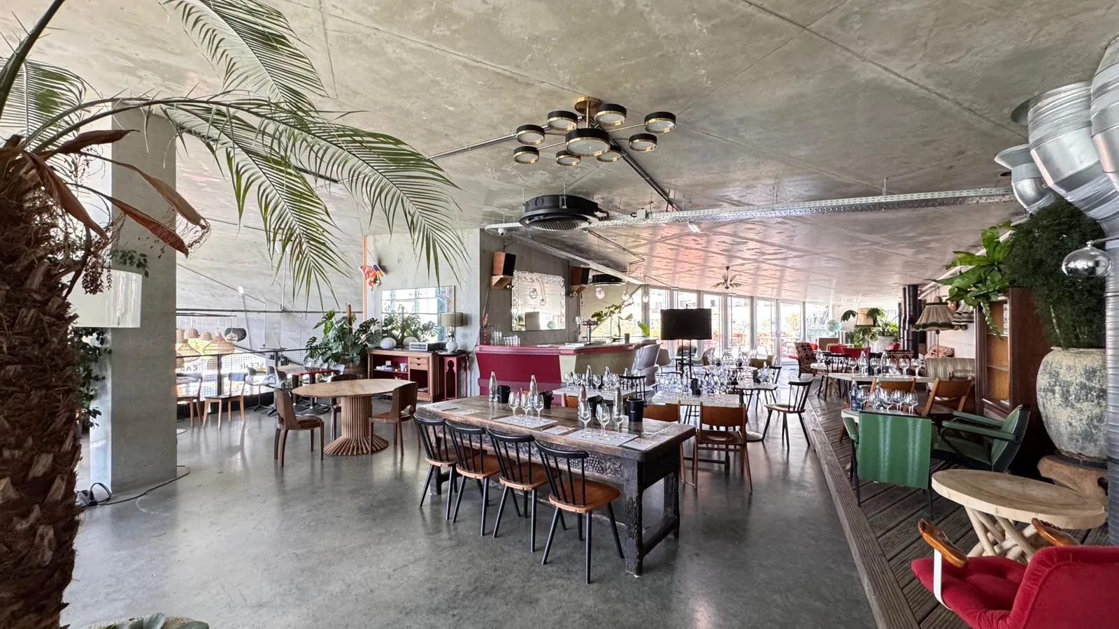 Spacious interior of a restaurant or event venue in Bordeaux featuring green plants, set tables, wooden and metal chairs, exposed concrete ceiling, and large windows.