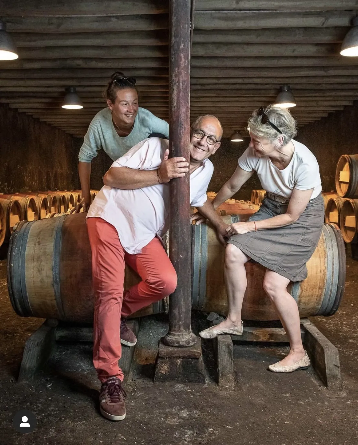 Three smiling people pose in front of wine barrels in a cellar at Château Baudan in Bordeaux, with one person hugging a central wooden pillar.