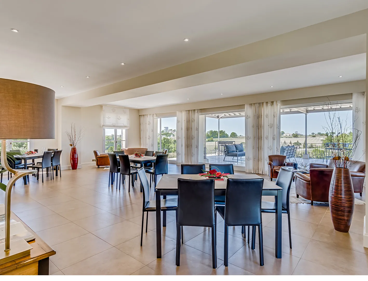 Spacious lounge and dining area at Château Tour Saint Christophe in Bordeaux, featuring modern black tables, brown leather chairs, large floor-to-ceiling windows opening onto a panoramic terrace, beamed ceiling, and contemporary decor.