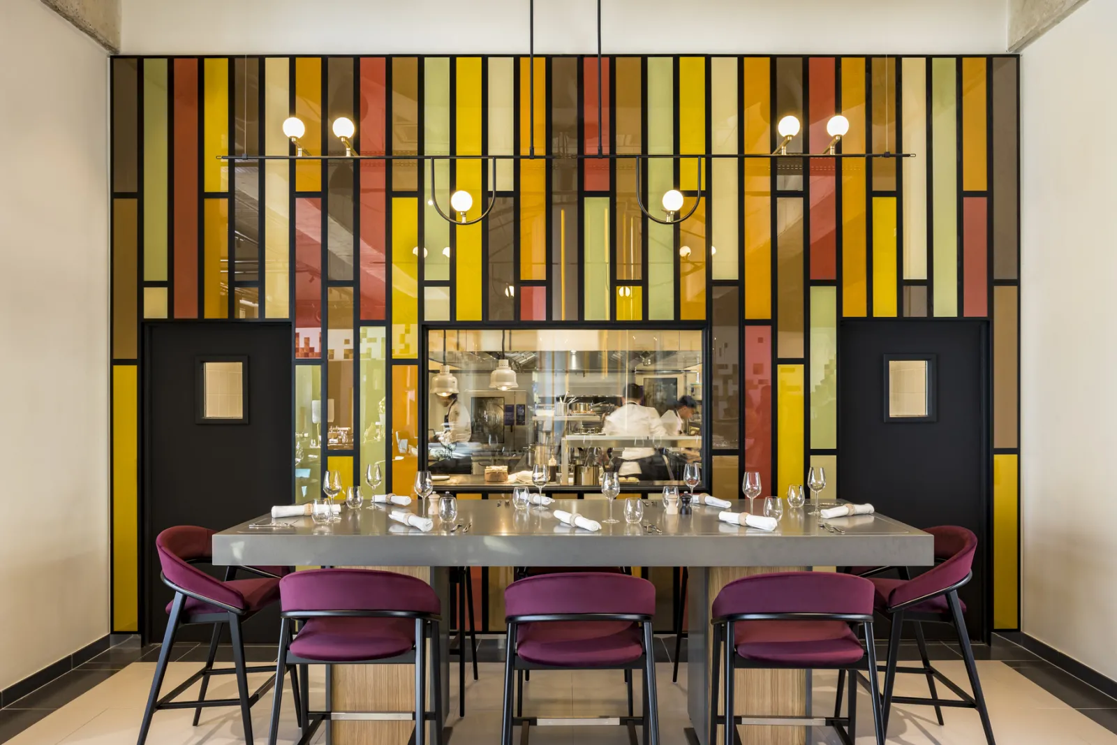 Le Bada restaurant dining area in Bordeaux featuring a grey marble counter, burgundy chairs, open kitchen view through glass, and geometric stained glass backdrop in yellow, ochre, red and black tones.