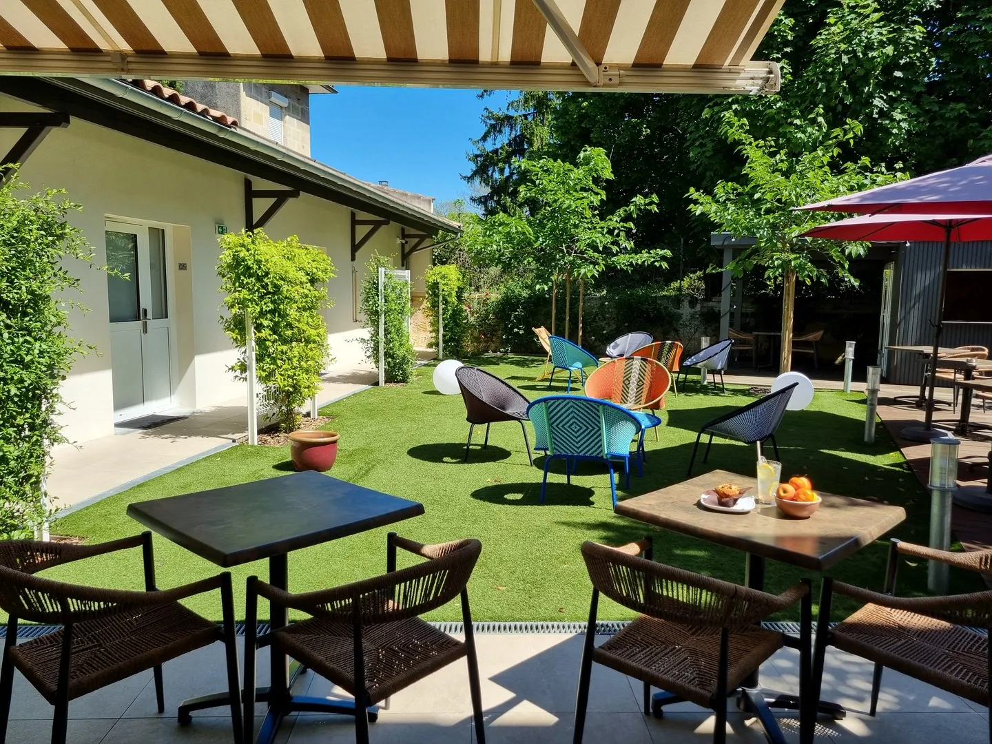 Sunny hotel terrace with colorful seating, dining tables, loungers, green lawn, umbrellas and white building in the background in Bordeaux