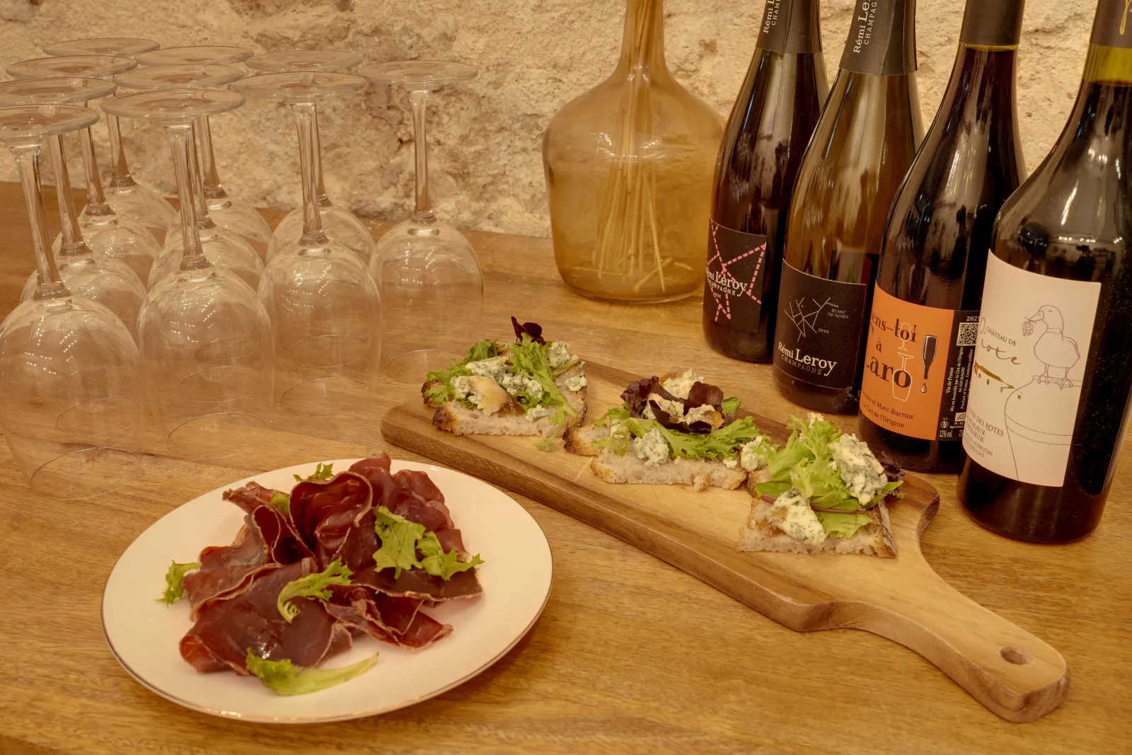 Wine tasting table in Bordeaux with empty glasses, wine bottles, cured meats and open-faced sandwiches with fresh cheese and greens against a stone wall background