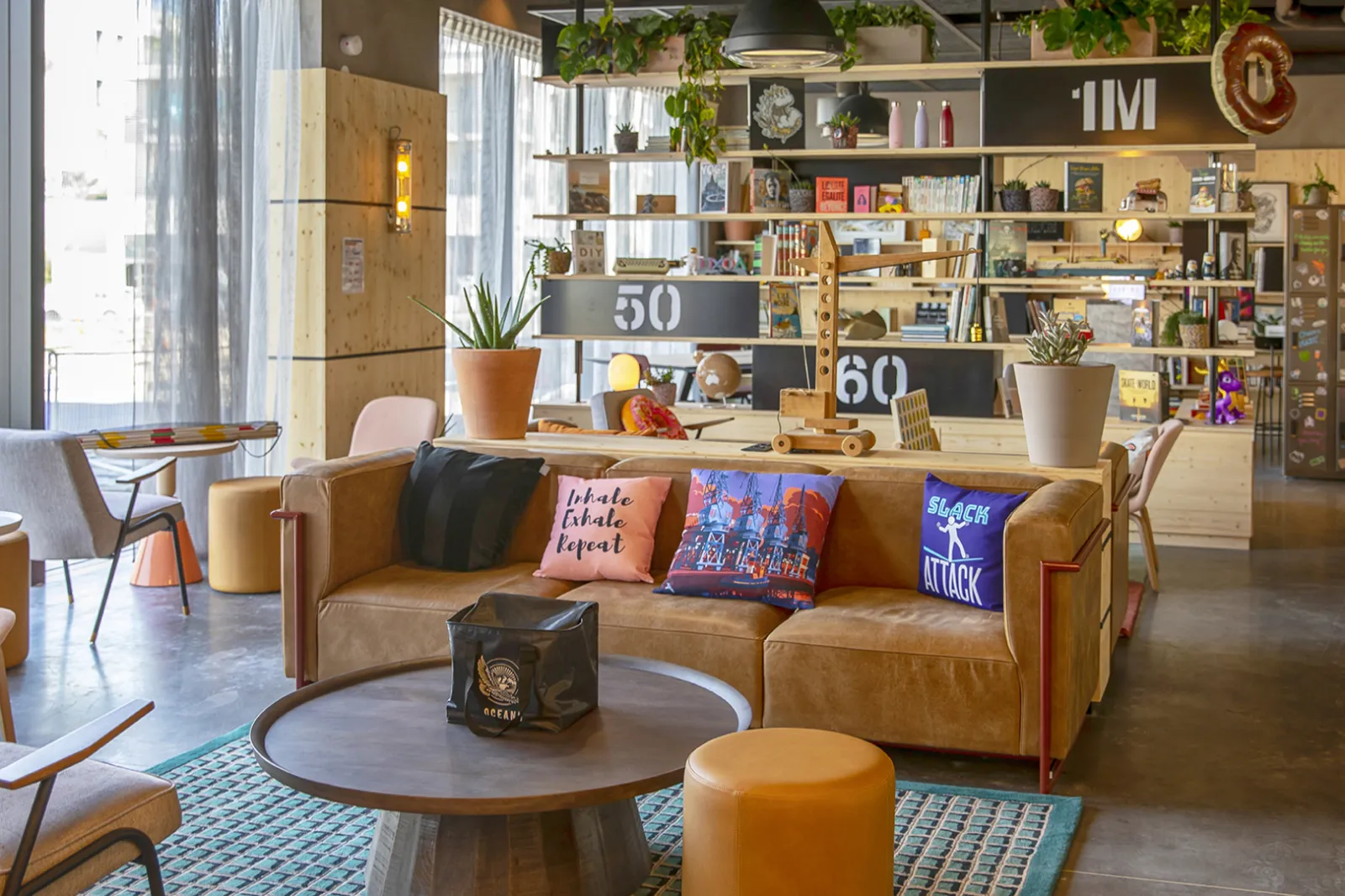 Modern and vibrant living space at Moxy Bordeaux featuring a brown leather sofa, round coffee table, wooden wall shelving with decorative items, and large windows overlooking the city.