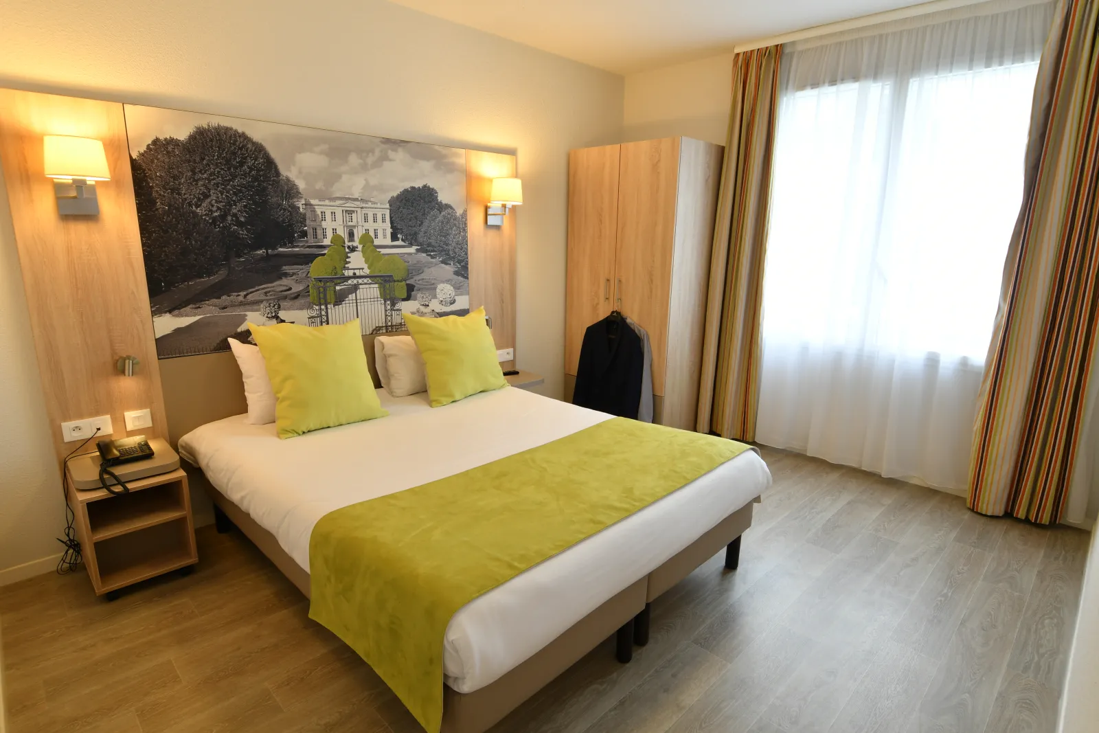 Modern bedroom in a Bordeaux aparthotel featuring a queen-size bed with white and lime-yellow linens, matching pillows, wooden nightstand, wall sconces, light wood wardrobe, classical mansion artwork above the bed, beige curtains, and floor-to-ceiling win