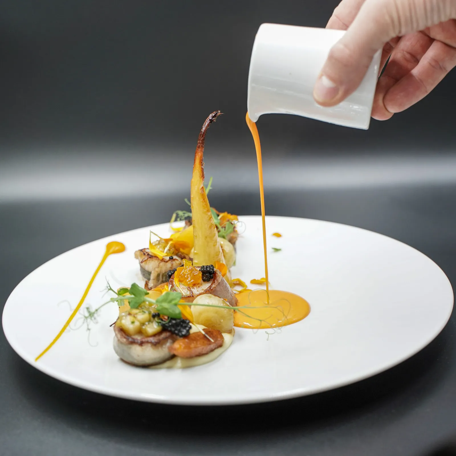 A chef pours a golden sauce over a gourmet dish featuring meat, vegetables and crispy elements artfully plated on white china.