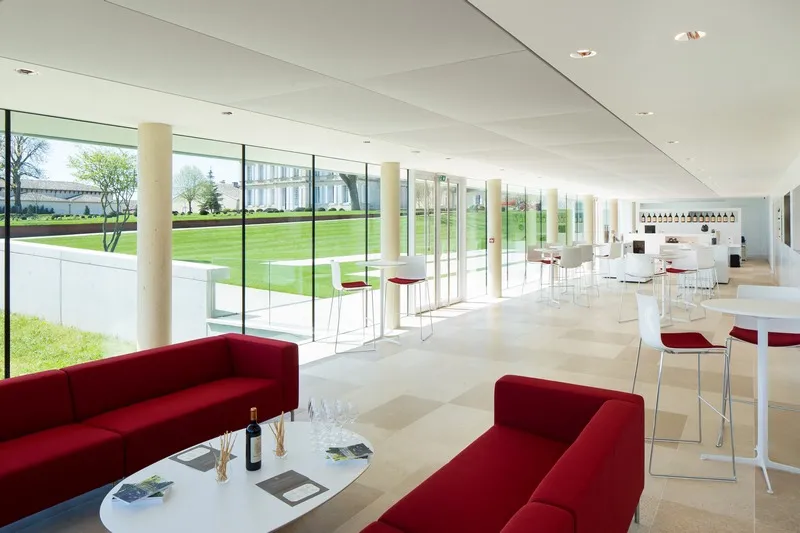 Modern bright reception hall featuring red sofas, white furniture, and expansive glass windows overlooking the vineyards of Château Gruaud Larose in Bordeaux.
