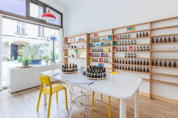 Bordeaux olfactory studio featuring a collection of wine bottles and luxury products displayed on wooden shelves, with a white table, yellow chairs, and large bay windows overlooking the city.
