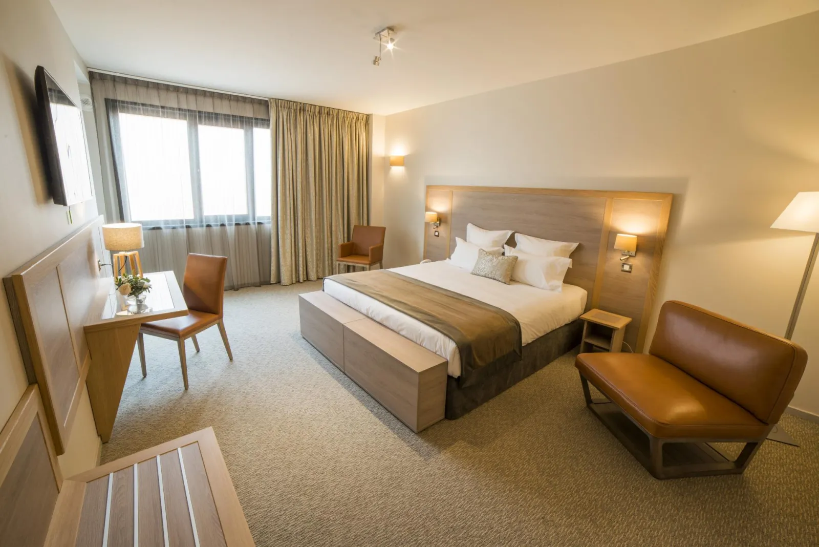 Elegant hotel room featuring a king-size bed, wooden furniture, brown leather sofa, desk, flat-screen TV and large windows with gold curtains at Vatel Hotel in Bordeaux