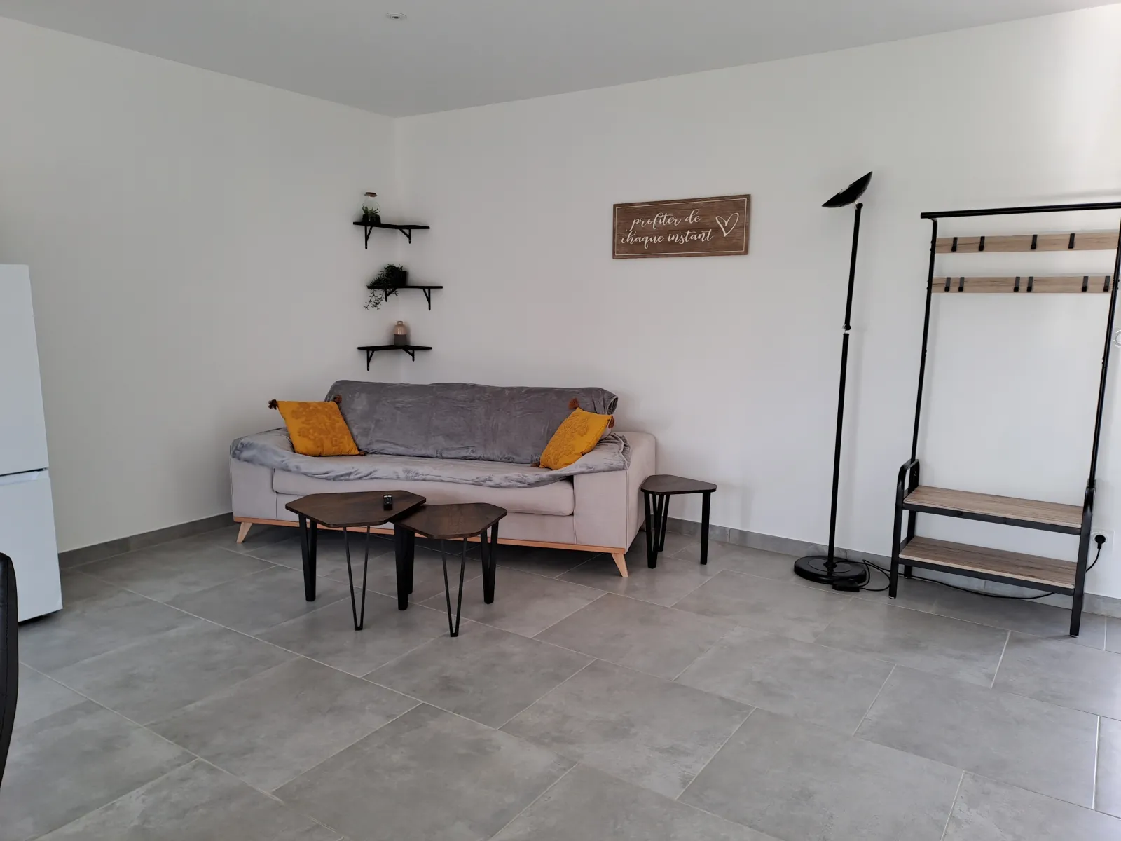 Modern interior of the Tanins guesthouse at Château Pierre de Montignac in Bordeaux, featuring a light gray sofa with orange cushions, black coffee table, wall shelves and a clothing rack on gray tiled flooring.