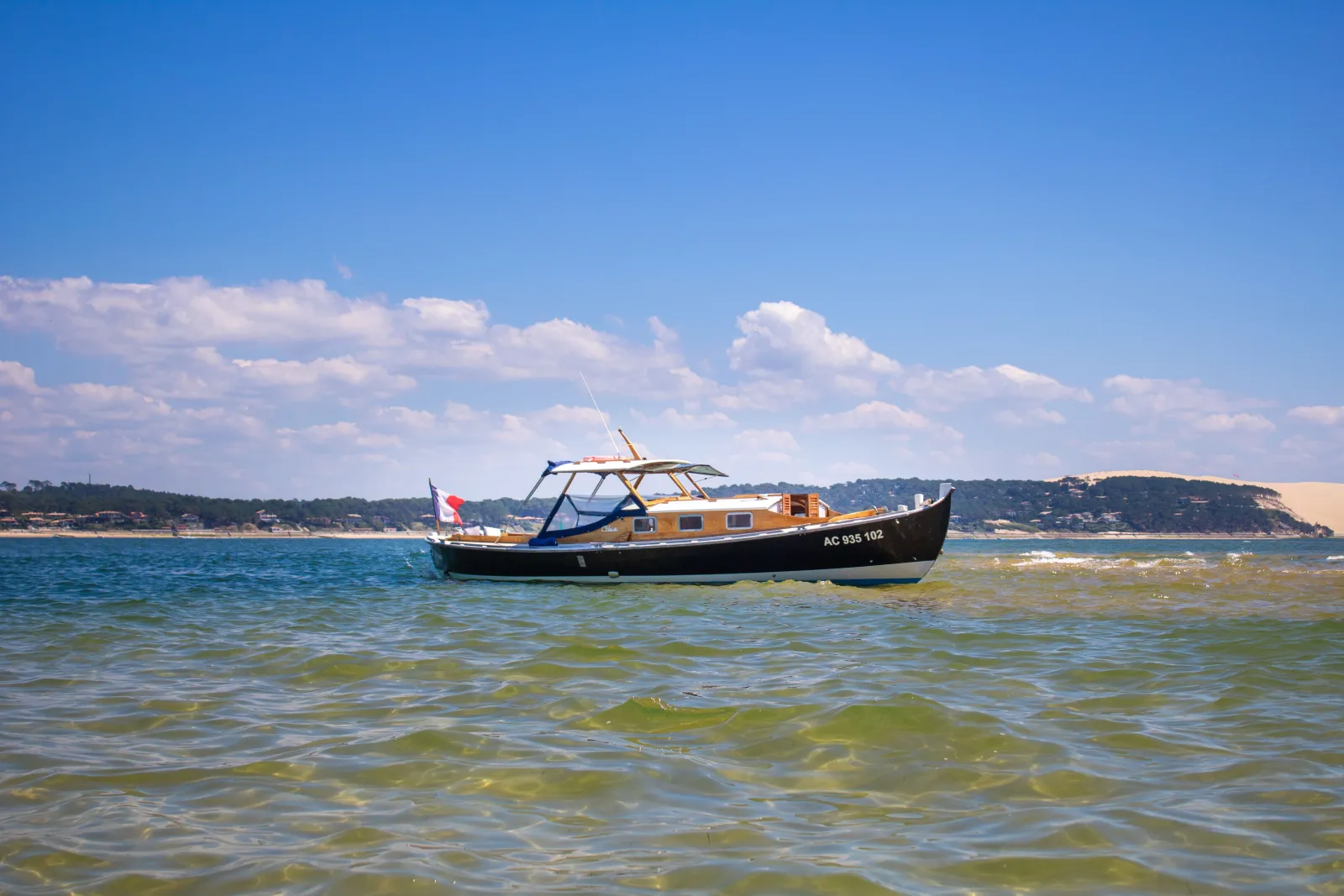 Altaïr pinasse, a traditional Bordeaux wooden boat painted in yellow and black, anchored in shallow waters with a French flag, against a blue sky and coastal landscape.