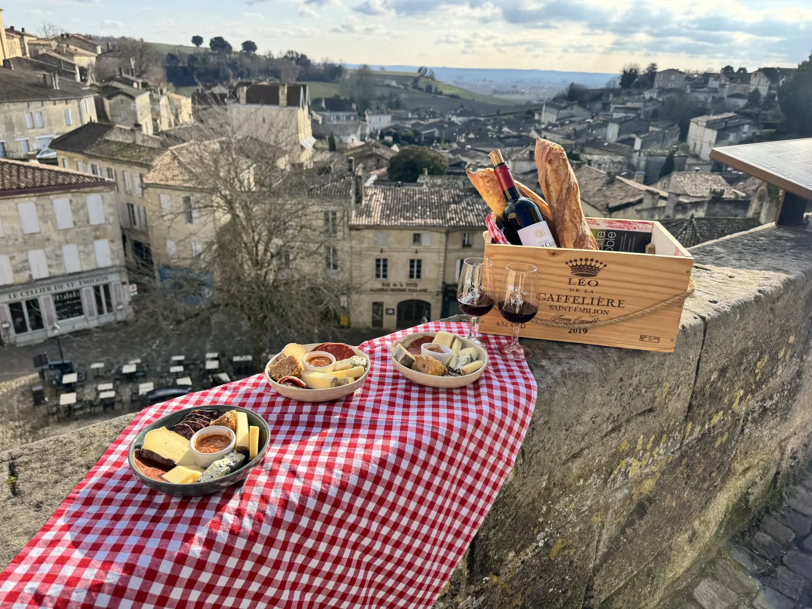 Gourmet picnic spread on a stone terrace overlooking a medieval French village. Red and white checkered tablecloth covers the table with charcuterie and cheese plates, red wine glasses, and a wooden wine box labeled Léo Caffelière.