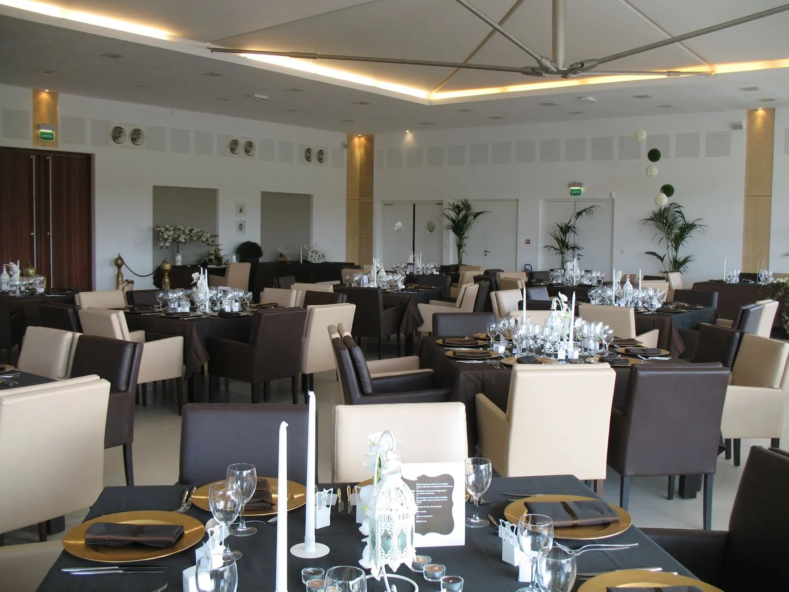 Elegant banquet hall at Château Luchey-Halde in Bordeaux featuring black and beige dining tables, matching upholstered chairs, place settings with glassware, decorative plants, and modern illuminated ceiling.