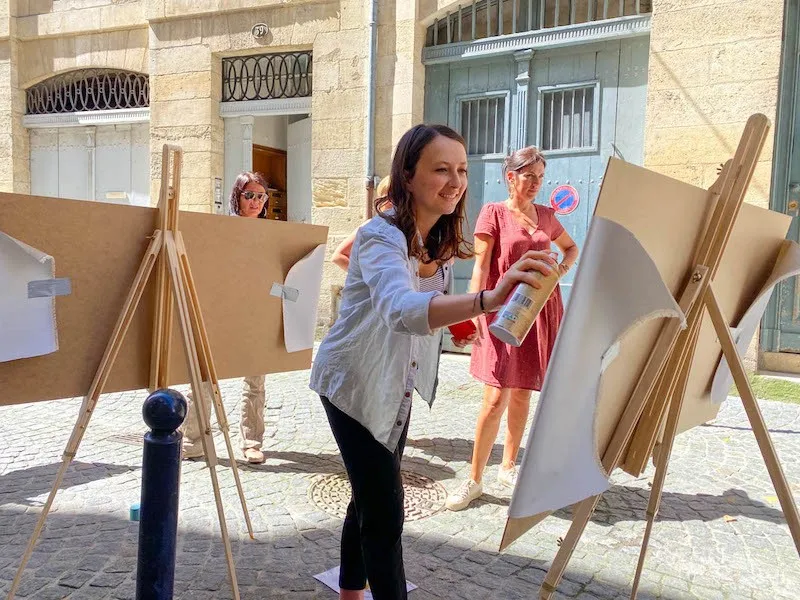 A woman paints on a cardboard easel in a paved courtyard in Bordeaux, surrounded by other participants in an outdoor art workshop.