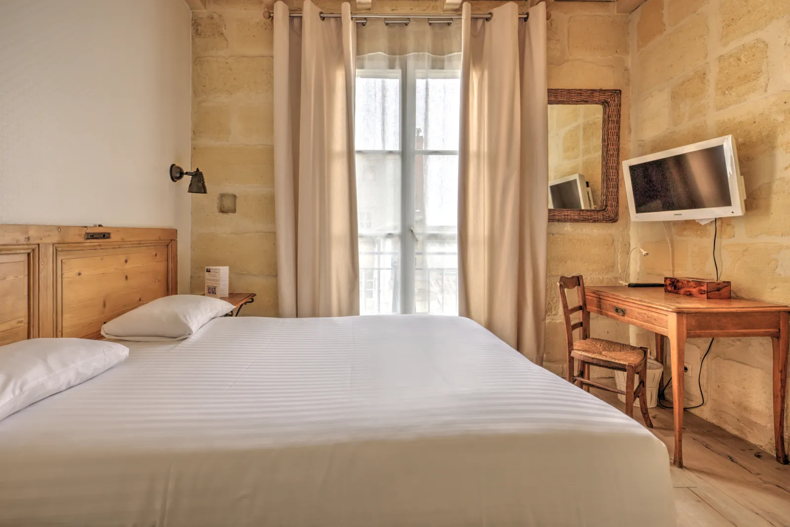 Elegant hotel room featuring a white bed, golden stone walls, light wood furniture, desk with chair, wall-mounted television, and tall windows with beige curtains in Bordeaux.