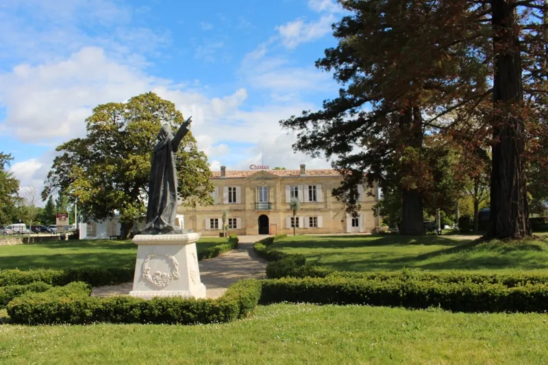 Château Marquis de Vauban in Bordeaux featuring an ochre stone façade surrounded by formal gardens, towering trees, and a contemporary sculpture in the foreground