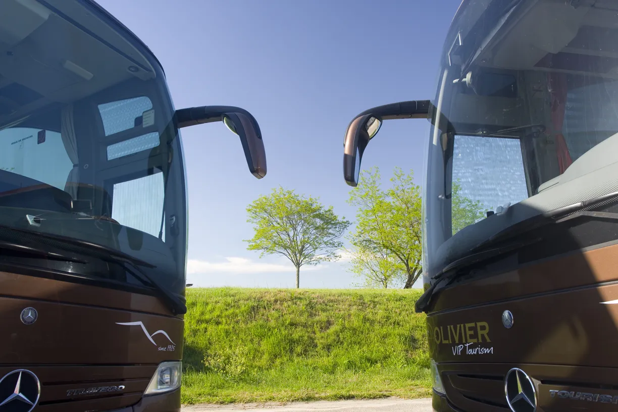 Two brown Mercedes-Benz coaches from Cars Olivier parked facing each other in a rural landscape with green fields and trees under blue sky