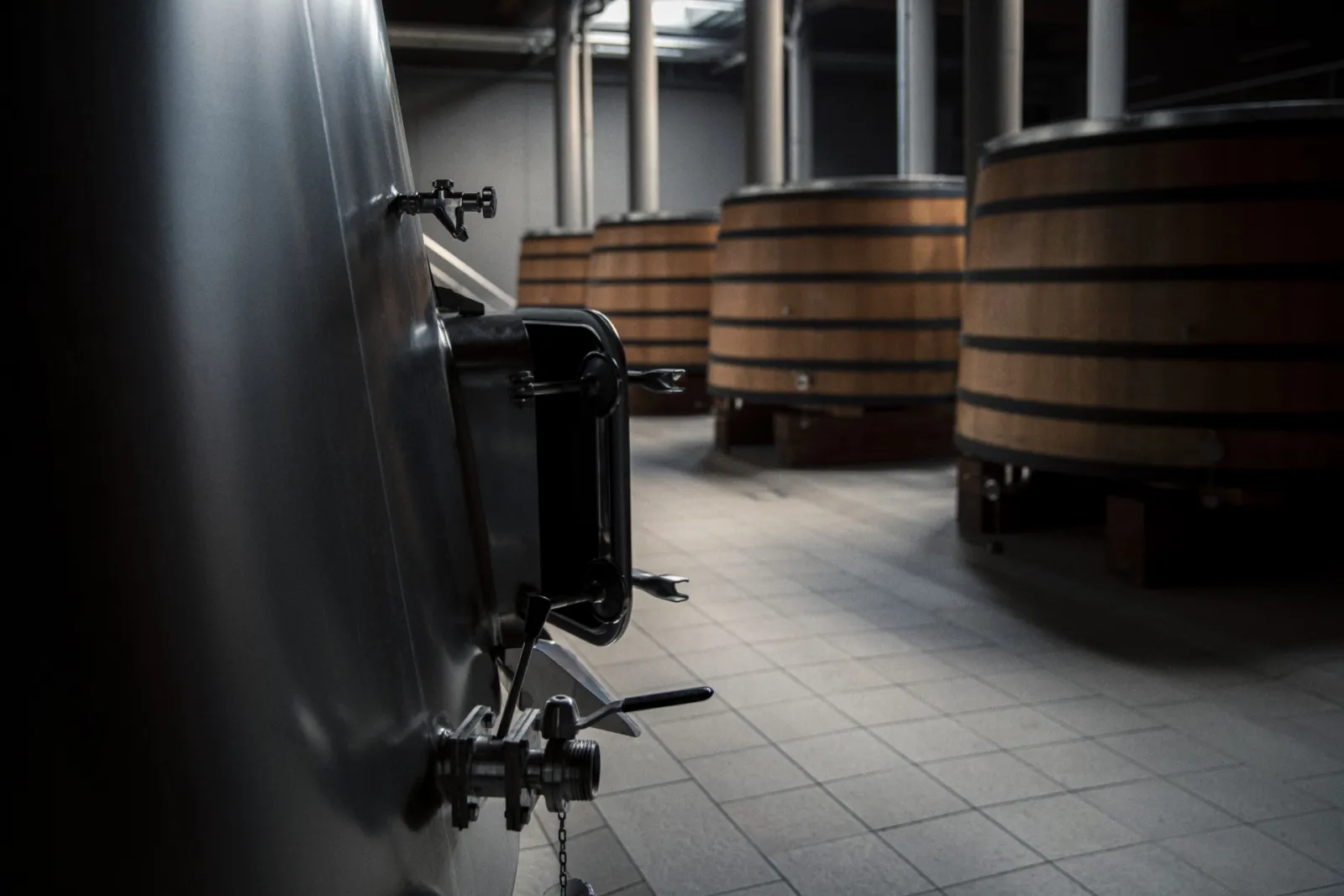 Wine cellar interior featuring a stainless steel barrel in the foreground and wooden oak barrels in the background, at Château de Fieuzal in Bordeaux.