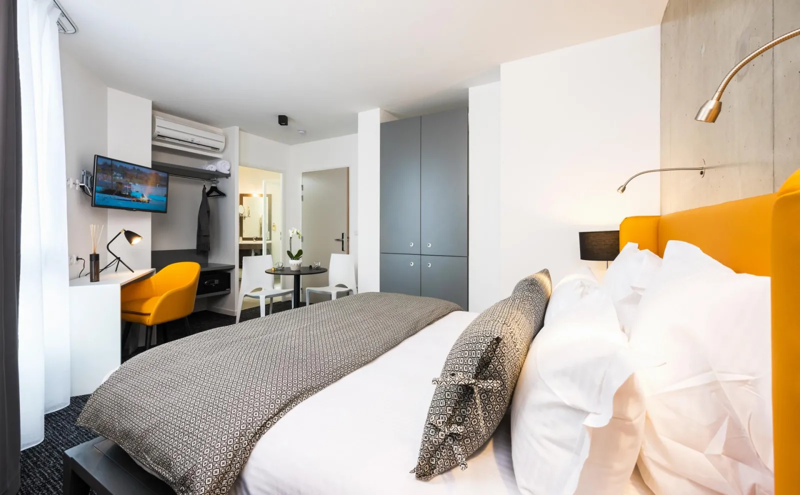 Modern hotel room featuring a double bed with orange headboard, yellow desk chair, wall-mounted TV, grey kitchenette, ensuite bathroom, and minimalist white and grey décor.