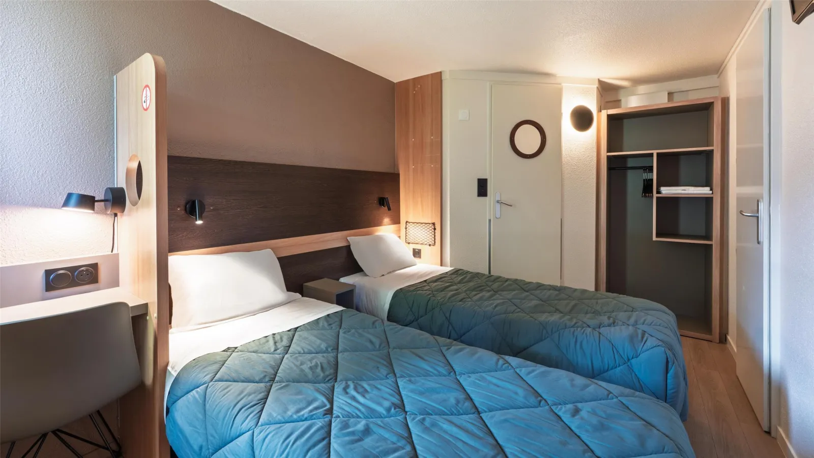 Modern hotel room at Première Classe Bordeaux Sud featuring two twin beds with blue bedding, brown headboard with integrated lighting, desk area, ensuite bathroom, and minimalist design.