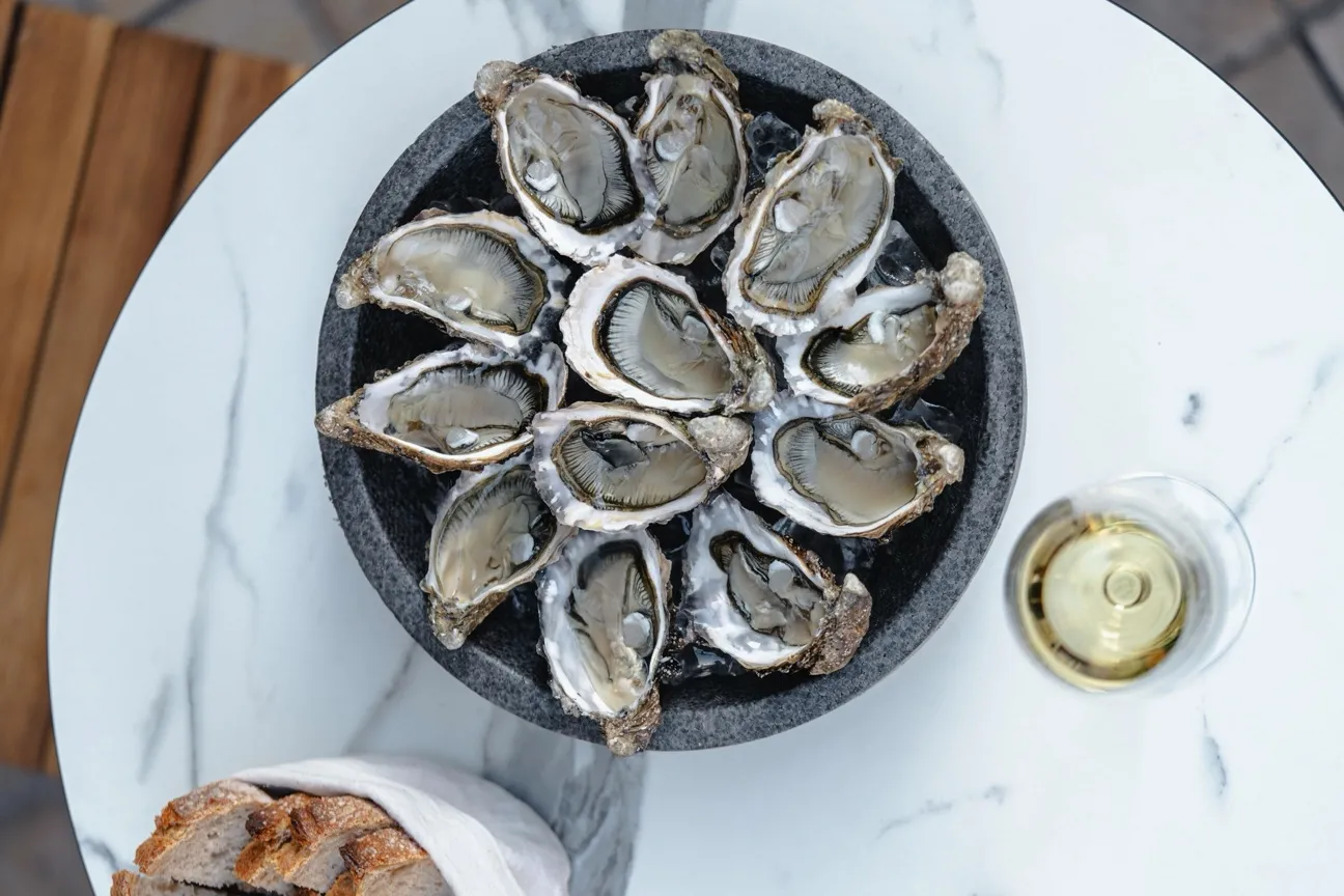 Plate of fresh oysters served on ice in a slate dish, accompanied by a glass of white wine and toasted bread