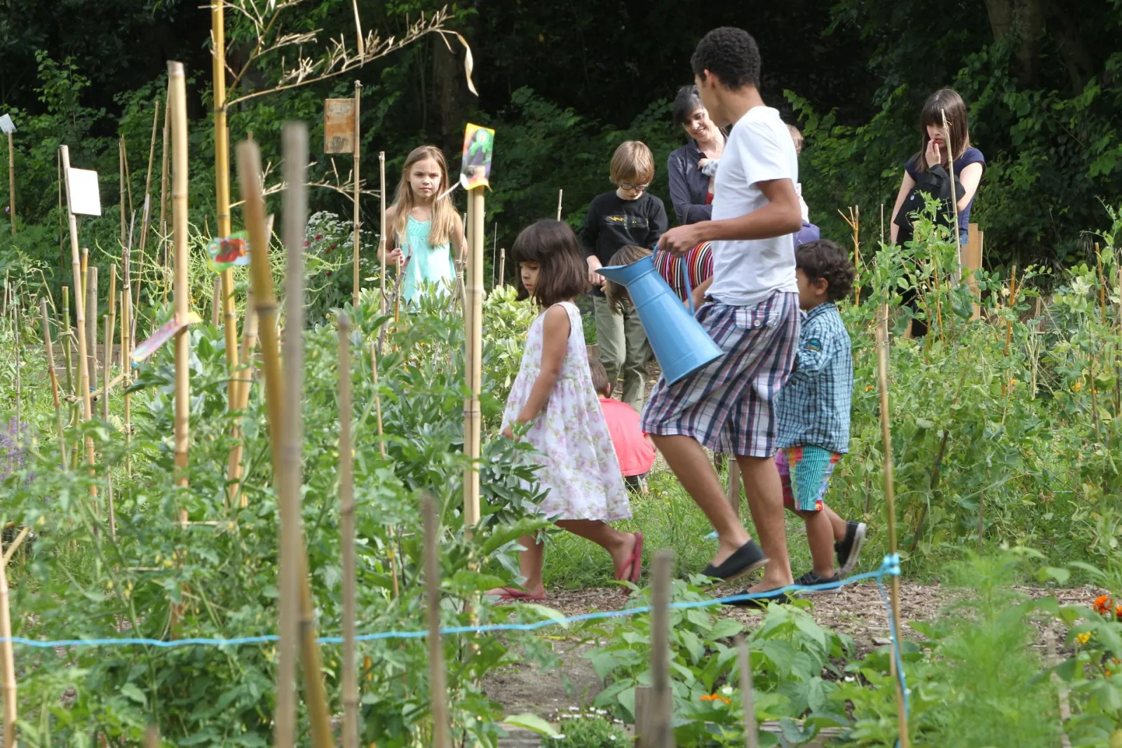 A group of children in a vegetable garden at Parc Rivière in Bordeaux, where a young adult demonstrates watering plants with a blue watering can.