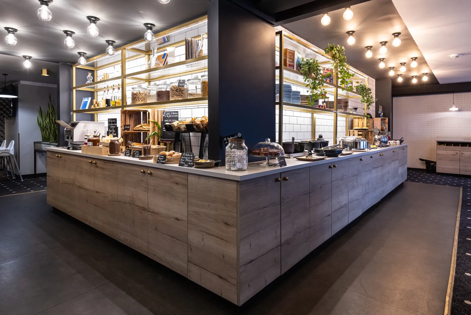 Modern café interior in Bordeaux featuring a light wood counter, illuminated golden shelving with products and décor, black ceiling with pendant lights, and polished concrete flooring.