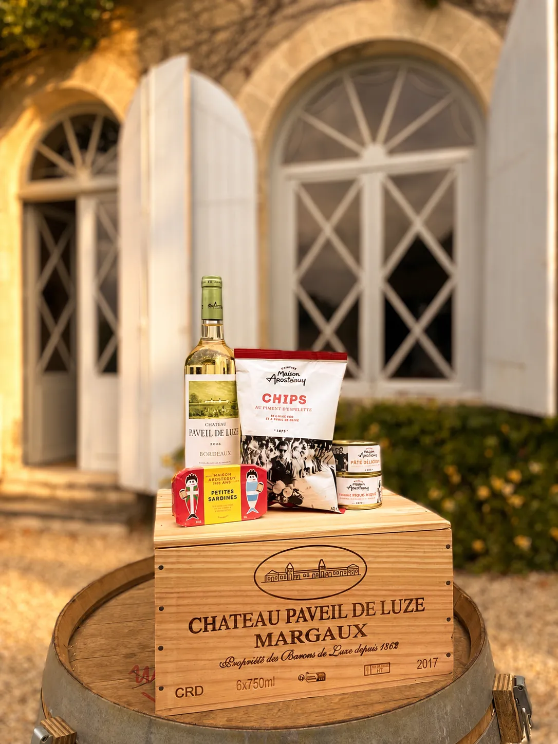 Gourmet basket from Château Paveil de Luze in Margaux featuring a white wine bottle, chips, crackers and regional products displayed on a wooden barrel in front of an illuminated Bordeaux villa