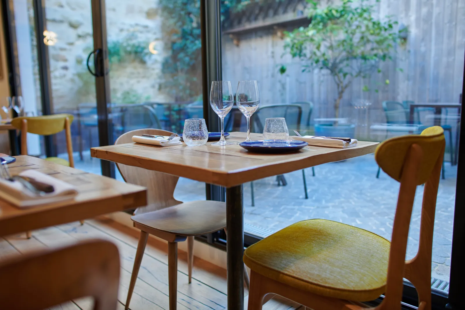 Elegant restaurant interior with set table, wine glasses and blue plates, light wood chairs, overlooking courtyard terrace in Bordeaux