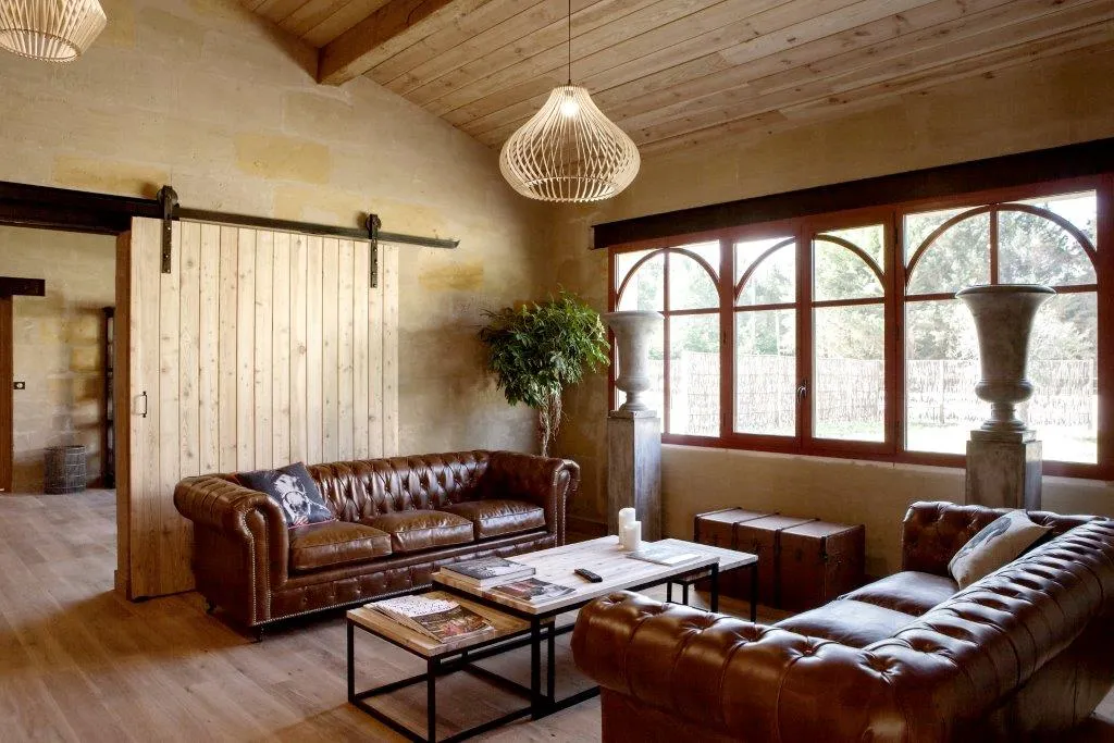 Spacious living room at Château Léognan featuring two brown leather Chesterfield sofas, wooden and metal coffee tables, arched French doors overlooking the grounds, exposed wooden ceiling beams and modern pendant lighting