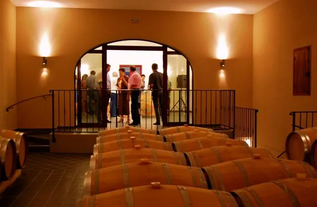 Interior of a wine cellar featuring wooden barrels in the foreground, an arched opening leading to a reception area with visitors, warm amber lighting.