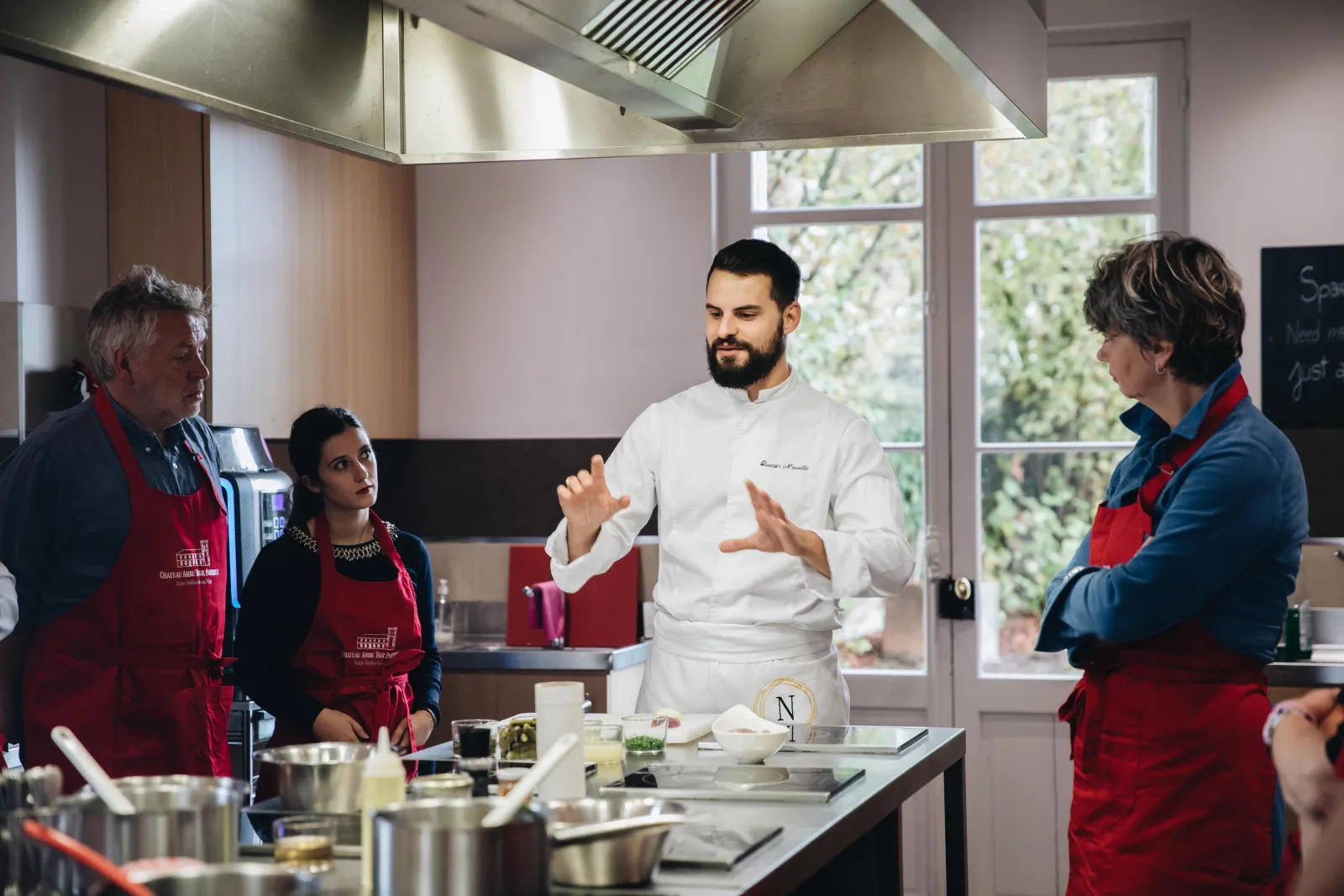 A chef in a white jacket gestures and explains to four participants wearing red aprons in a modern professional kitchen at Château Ambe Tour Pourret in Bordeaux.