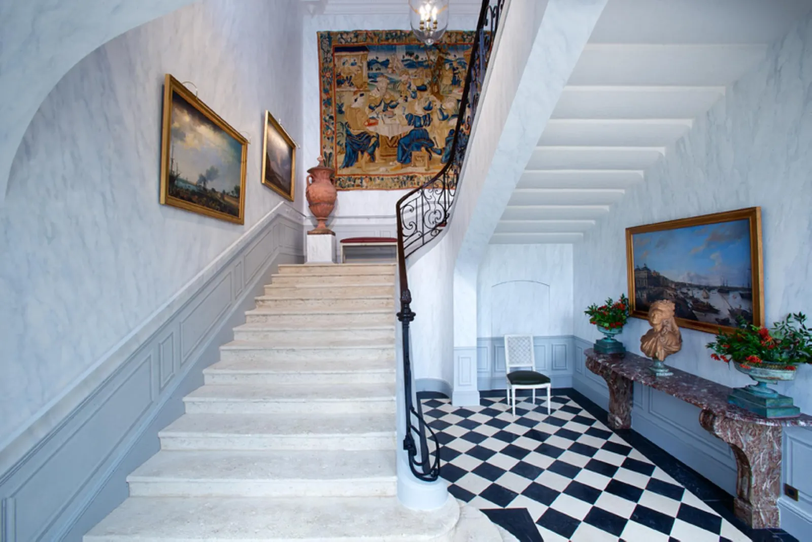 Elegant staircase in a Bordeaux castle featuring white steps, black and white checkered flooring, wrought iron railing, framed paintings, antique tapestry on ceiling, and classical décor with busts and vases.
