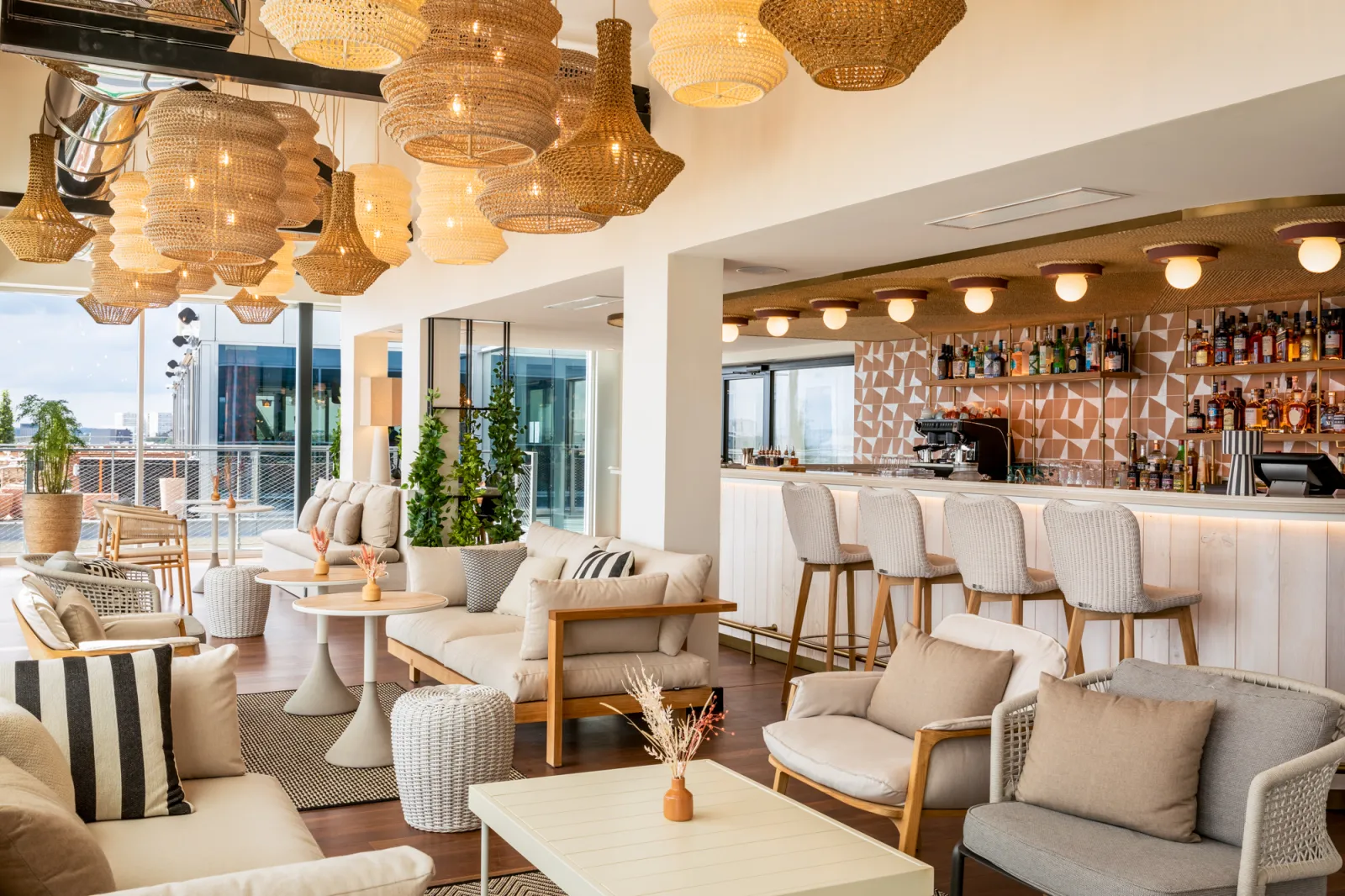 Sophisticated lounge bar featuring cream and natural furnishings, woven pendant lights, bar counter in background, panoramic terrace overlooking Bordeaux through large windows