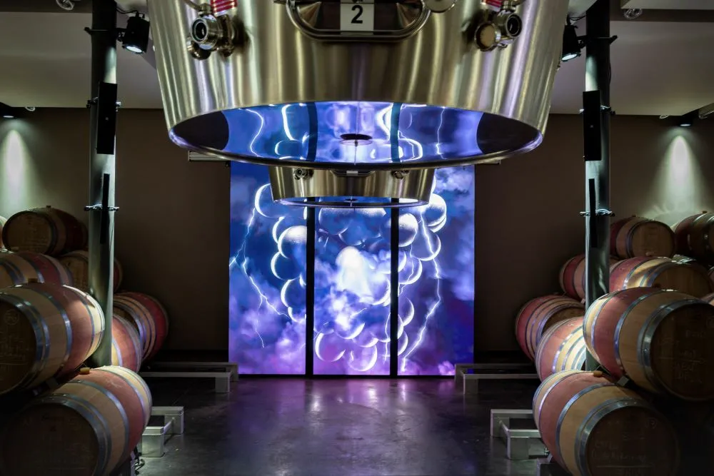 Modern winery facility at Château La Fleur de Boüard in Bordeaux, featuring oak barrels on either side and a large digital screen displaying an illuminated grape cluster pattern in blue and purple.