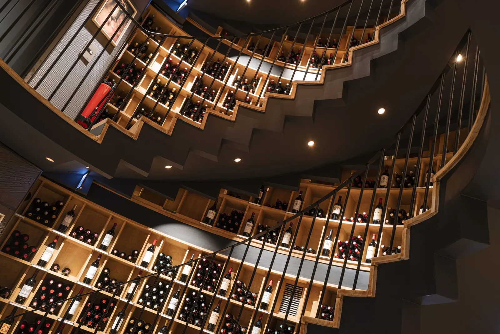Overhead view of a modern wine cellar with wooden shelves arranged in a spiral pattern, displaying numerous illuminated wine bottles against dark walls with a metal staircase.