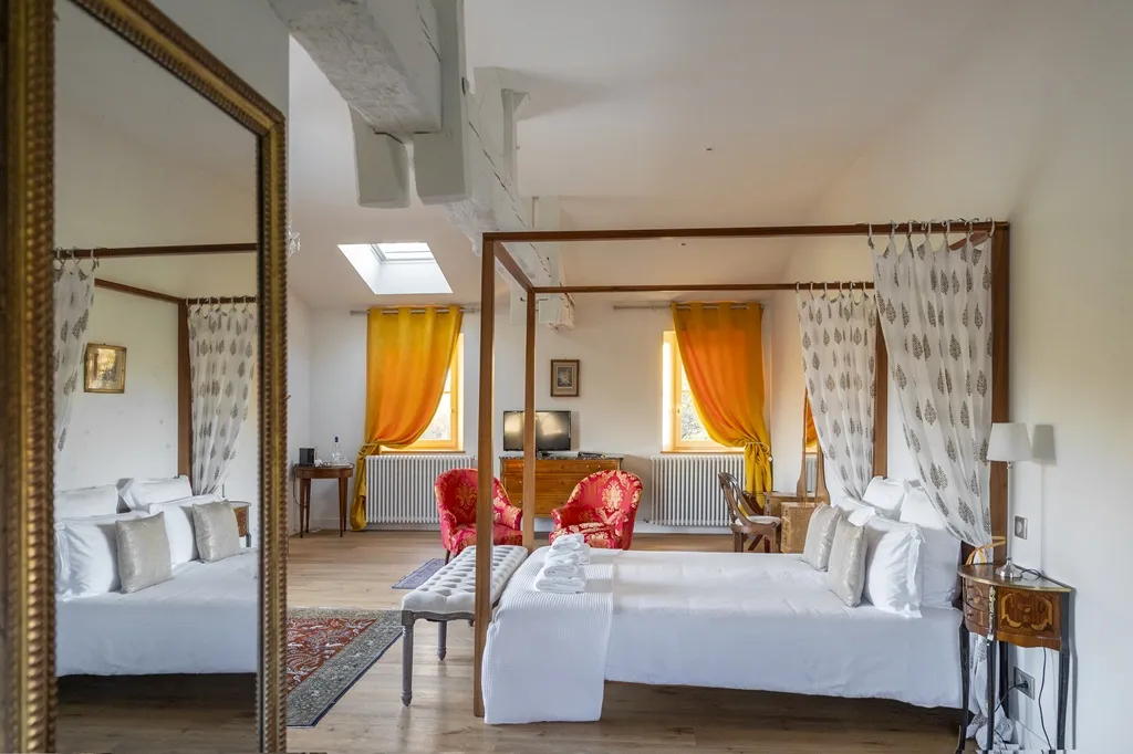 Luxury hotel bedroom featuring two wooden four-poster beds, orange and white curtains, gilded mirror, red armchairs, and natural light from skylights
