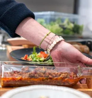 Hand of a person wearing gold bracelets pouring a spicy sauce into a glass container, with vegetables and herbs in the background in a kitchen.
