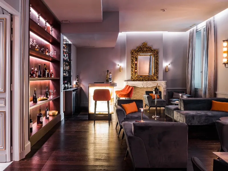 Elegant lounge at Hôtel Villas Foch in Bordeaux featuring illuminated bar, modern gray and orange velvet furniture, gold framed mirror, and ambient pink lighting
