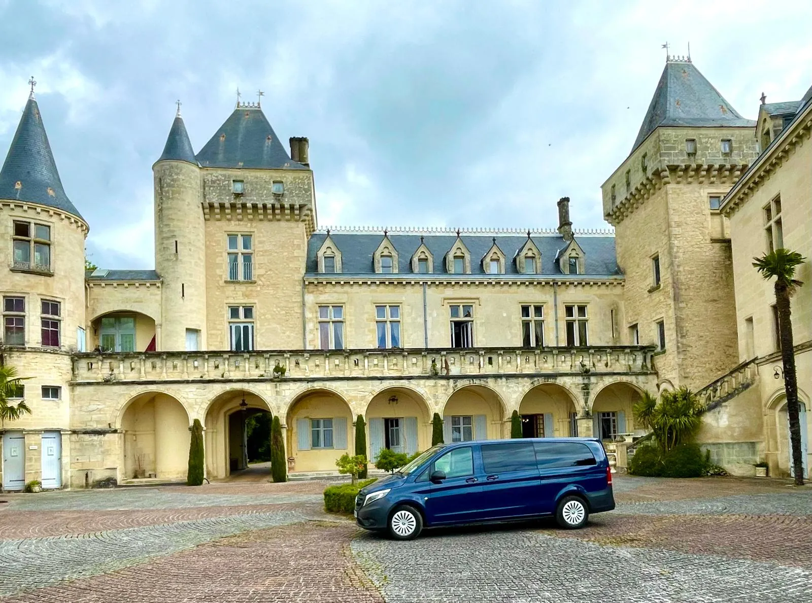 Château de la Malromé in Bordeaux featuring pointed turrets, stone facade, vaulted arcades and a blue minivan parked in the brick courtyard