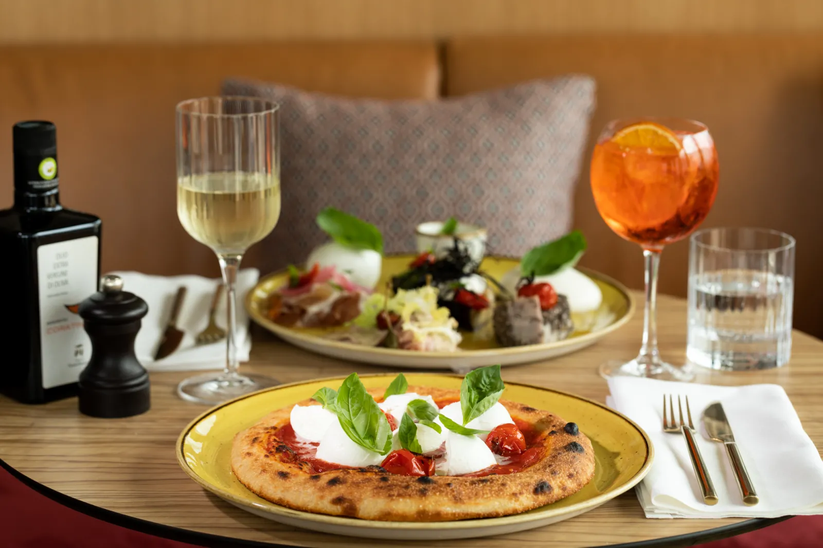 Elegant restaurant table featuring two gourmet pizzas topped with fresh mozzarella and basil, paired with glasses of white wine and an orange cocktail, set in a refined ambiance with beige sofa in the background