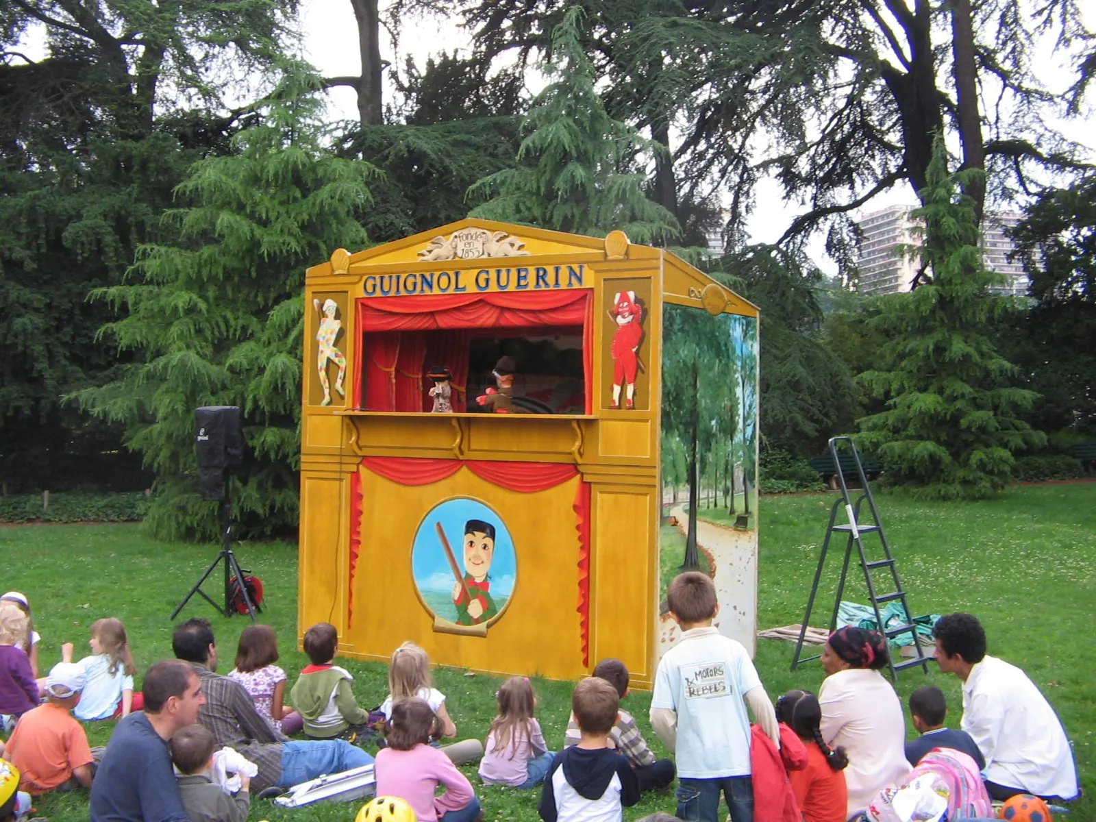 Guignol Guérin puppet theater in Bordeaux, a yellow and red wooden structure with children seated on grass in a park watching the show.