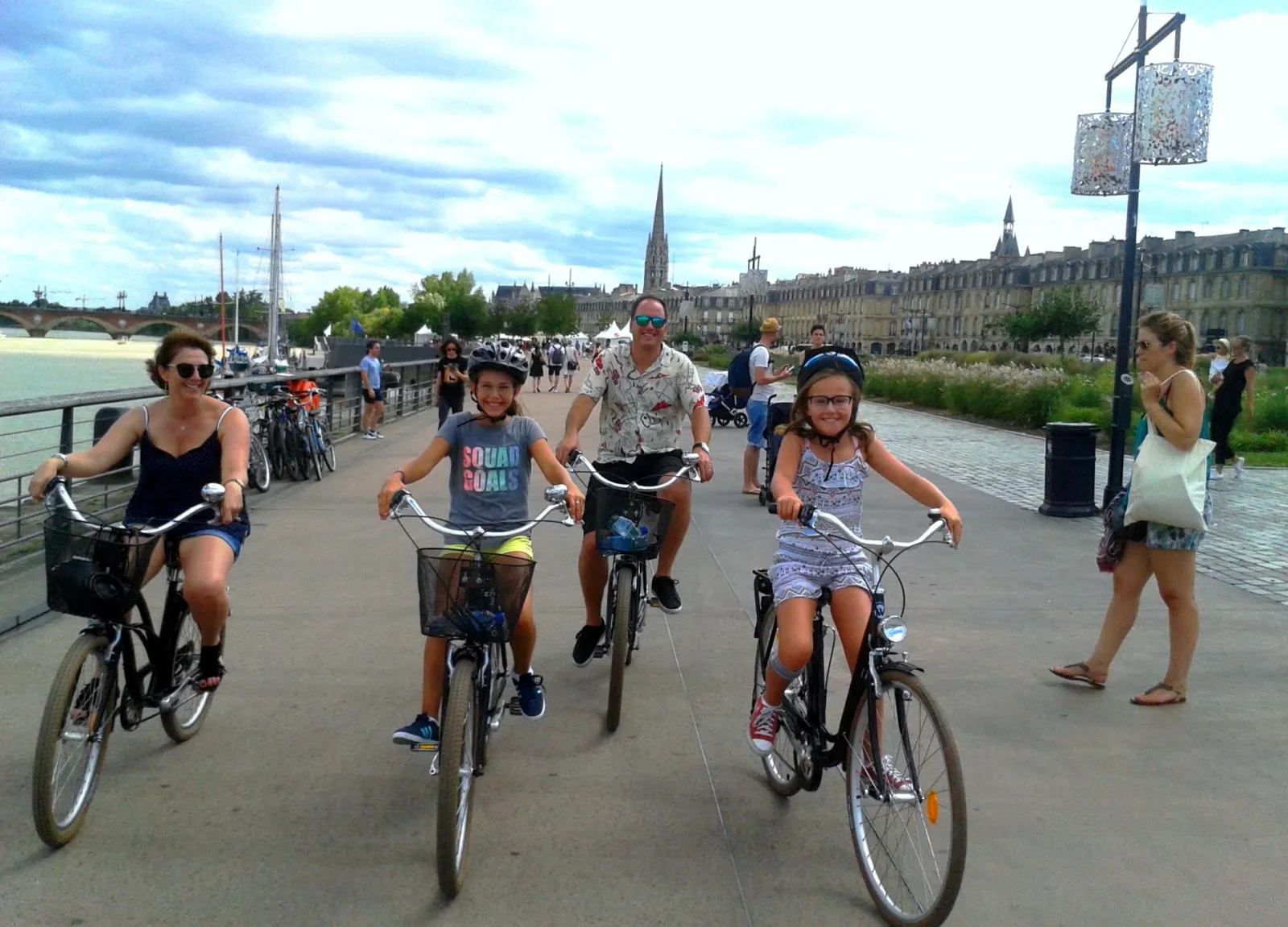 Family cycling along the Garonne riverside promenade in Bordeaux, with children and adults riding bikes on a paved path, historic city buildings and church spires visible in the background.