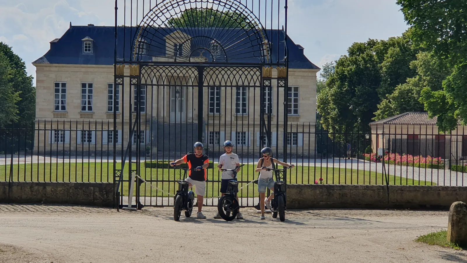 Three children on bicycles in front of ornamental gates of an elegant classical building with blue mansard roof and stone facade in Bordeaux.