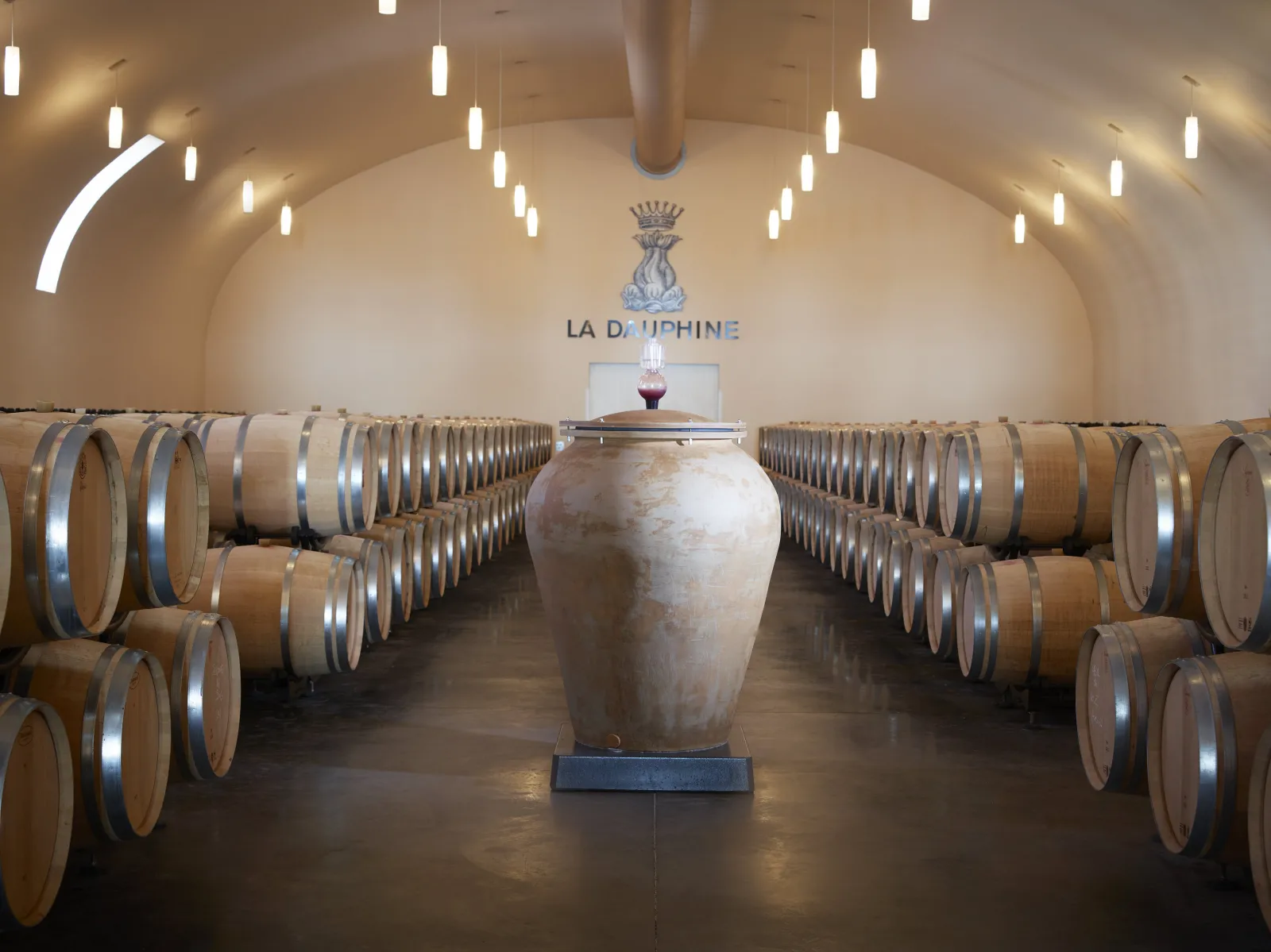 Wine cellar of Château de La Dauphine in Bordeaux featuring rows of oak barrels on both sides, a large stone amphora in the center, and the château's logo on the vaulted ceiling.
