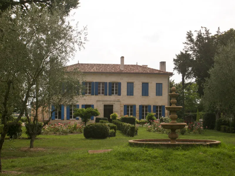Elegant neoclassical villa with beige façade and blue shutters, surrounded by a landscaped park featuring an ornamental fountain, trees and lush lawn.