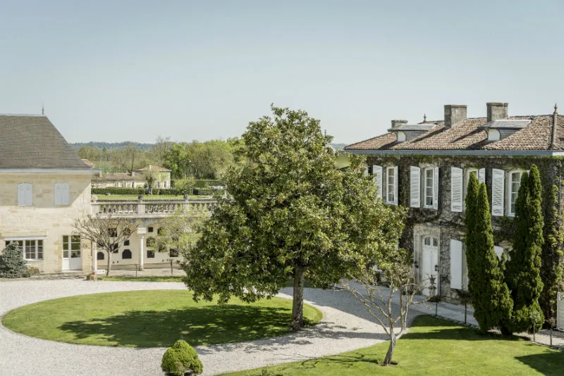 Aerial view of Château Prieuré-Lichine in Bordeaux featuring white stone buildings with tiled roofs, a large tree in the courtyard, gravel pathways, and manicured gardens with vineyards in the background.