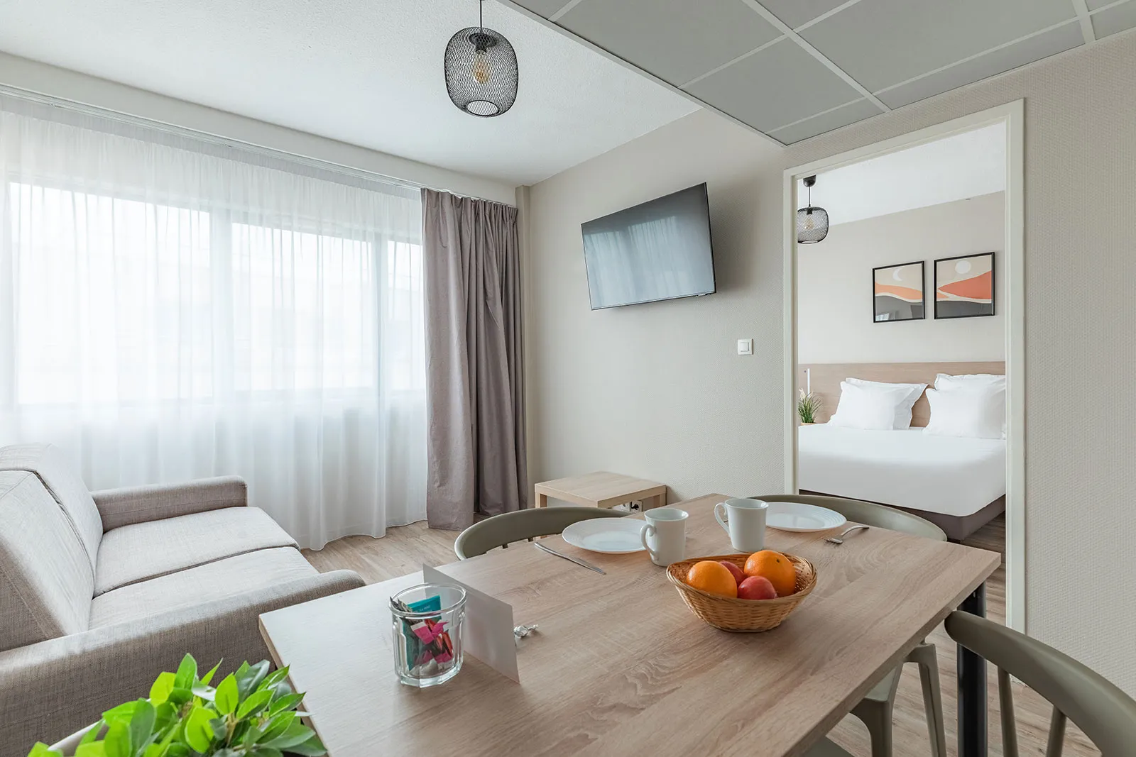 Spacious suite in a serviced apartment in Bordeaux featuring living area, dining table, gray sofa, bright windows and bedroom in background