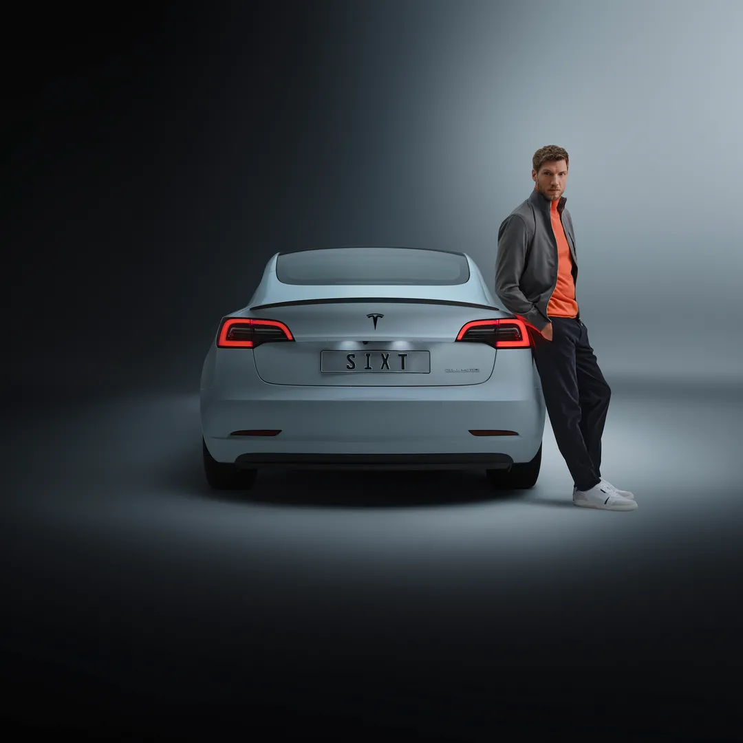 A man in grey jacket and orange shirt leaning against a white Tesla with SIXT license plate, on neutral grey background